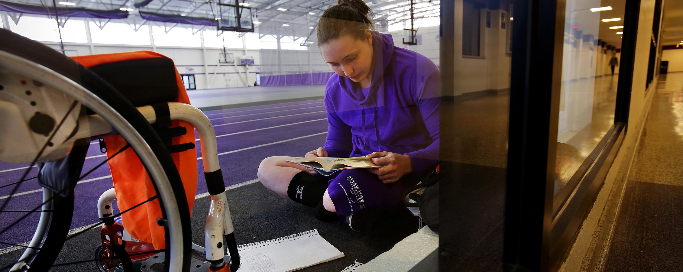 A person sits on the floor in front of their wheelchair as they read a book.