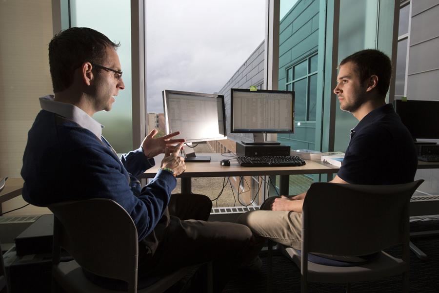 A student and a faculty member have a discussion at a table by two computers.