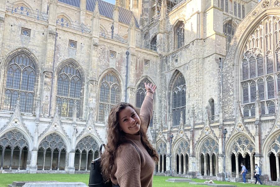 A young woman stands in a courtyard and gestures up toward a European cathedral.